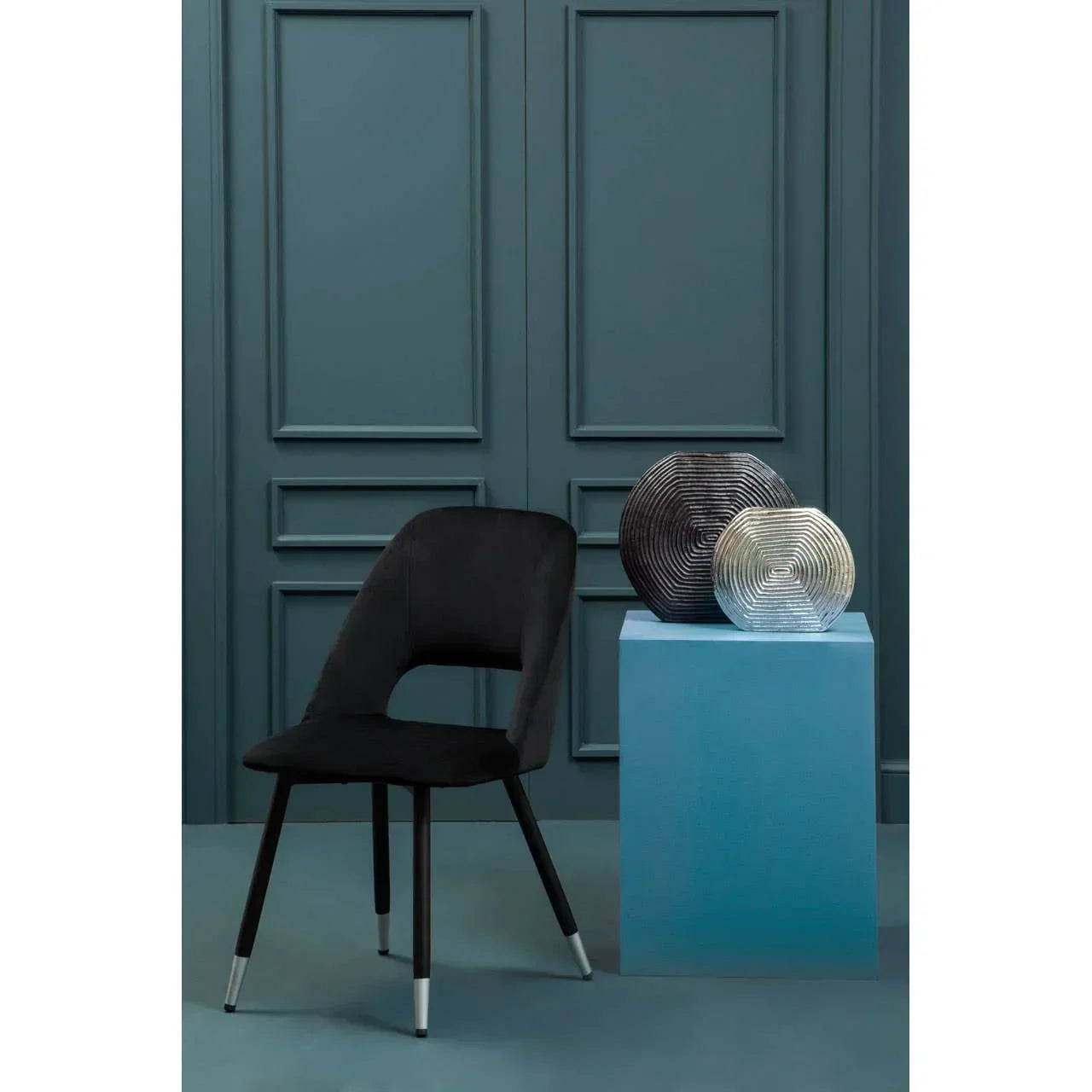 Black velvet dining chair with metal legs beside blue pedestal and two modern vases