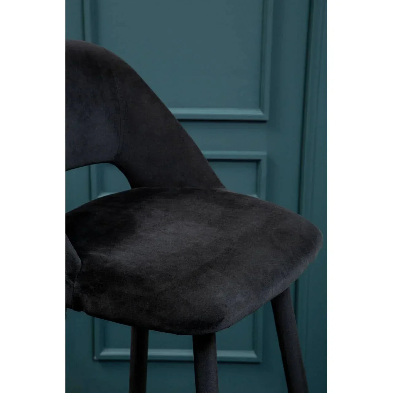Black velvet bar stool with curved backrest and tapered legs against teal panelled wall