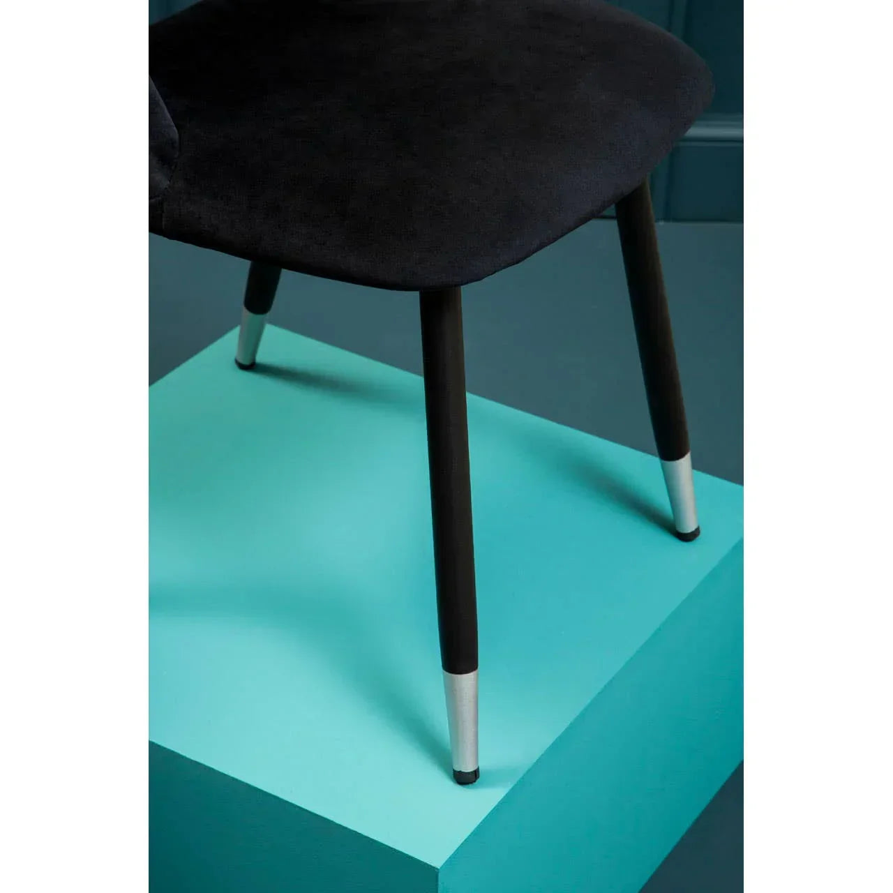 Black velvet dining chair with metal tipped wooden legs on blue platform