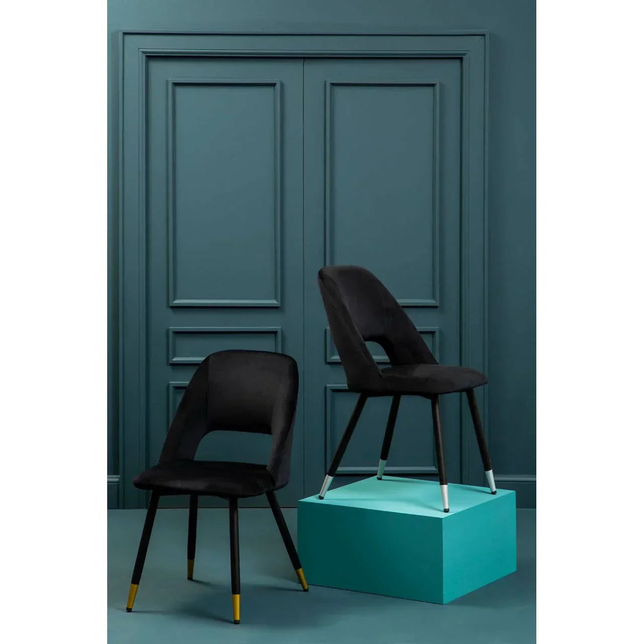 Pair of black velvet dining chairs with gold and silver leg caps against blue paneled wall