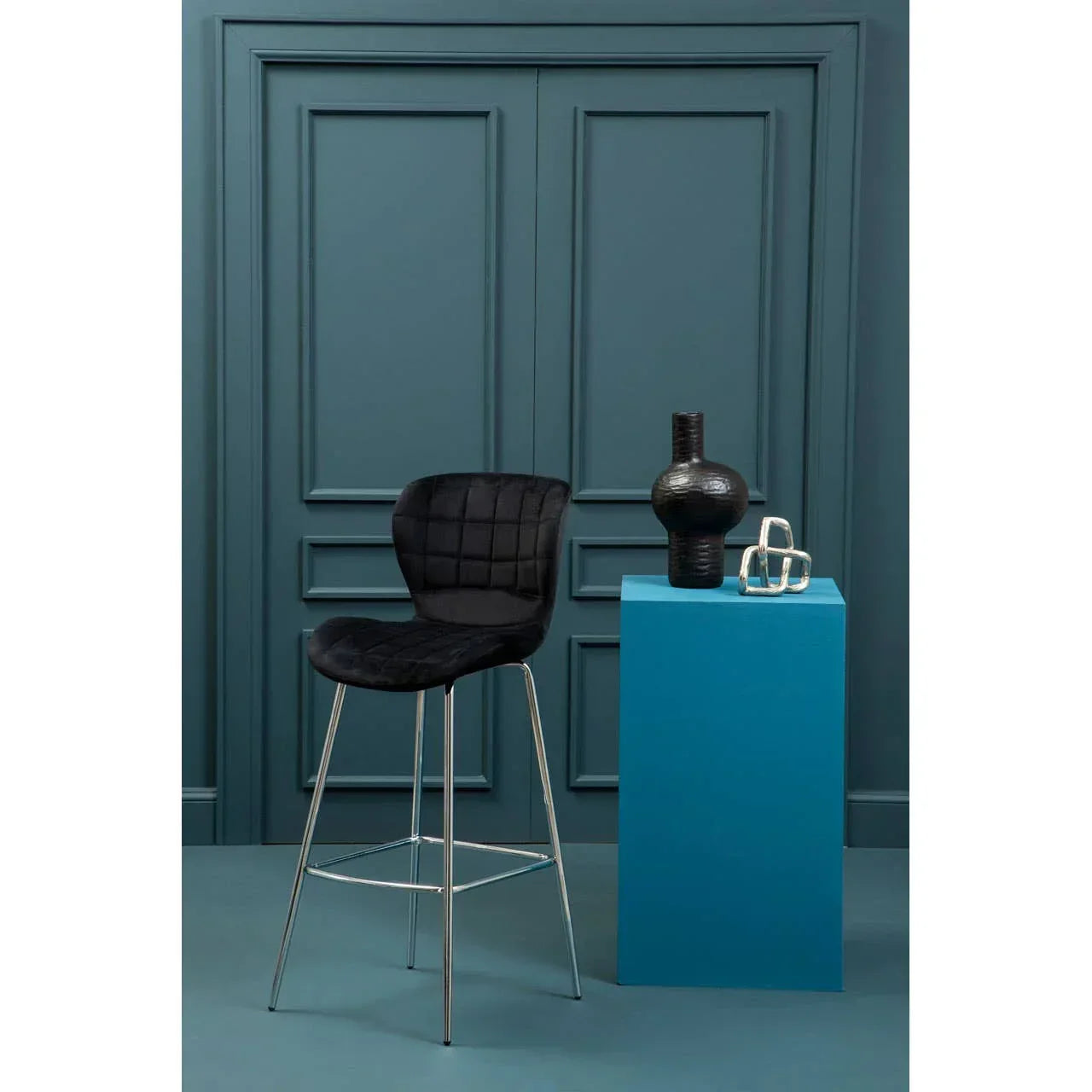 Black velvet bar stool with chrome legs beside blue pedestal and modern decor