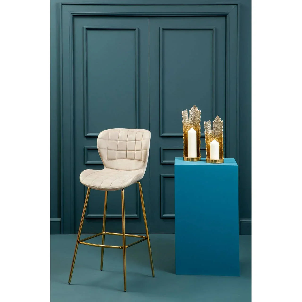 Cream bar stool with gold metal legs beside blue pedestal and decorative candles