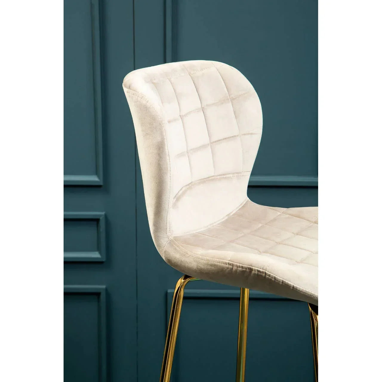 Beige velvet bar stool with gold metal legs and quilted seat detail against blue wall