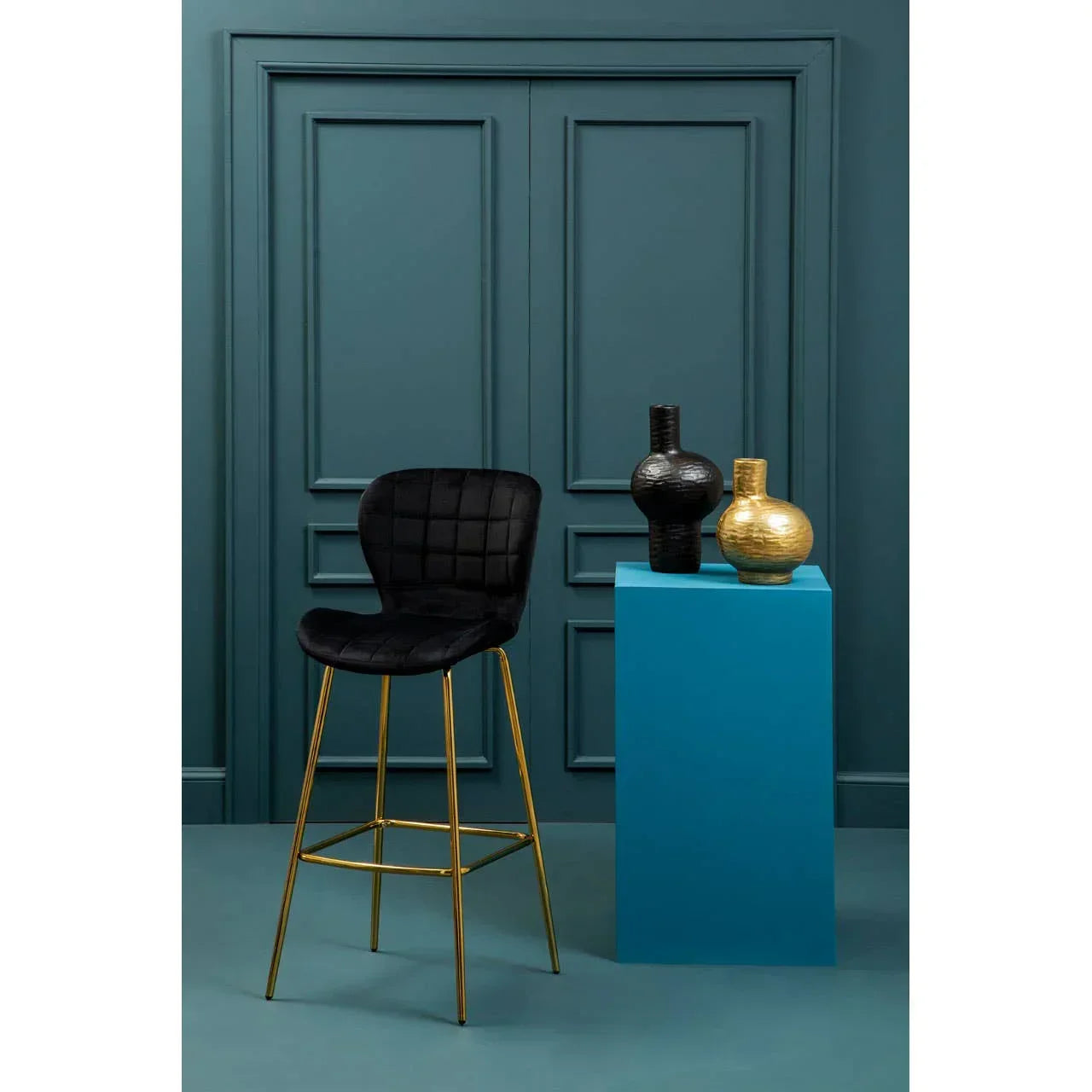 Black velvet bar stool with gold legs beside blue pedestal and decorative vases