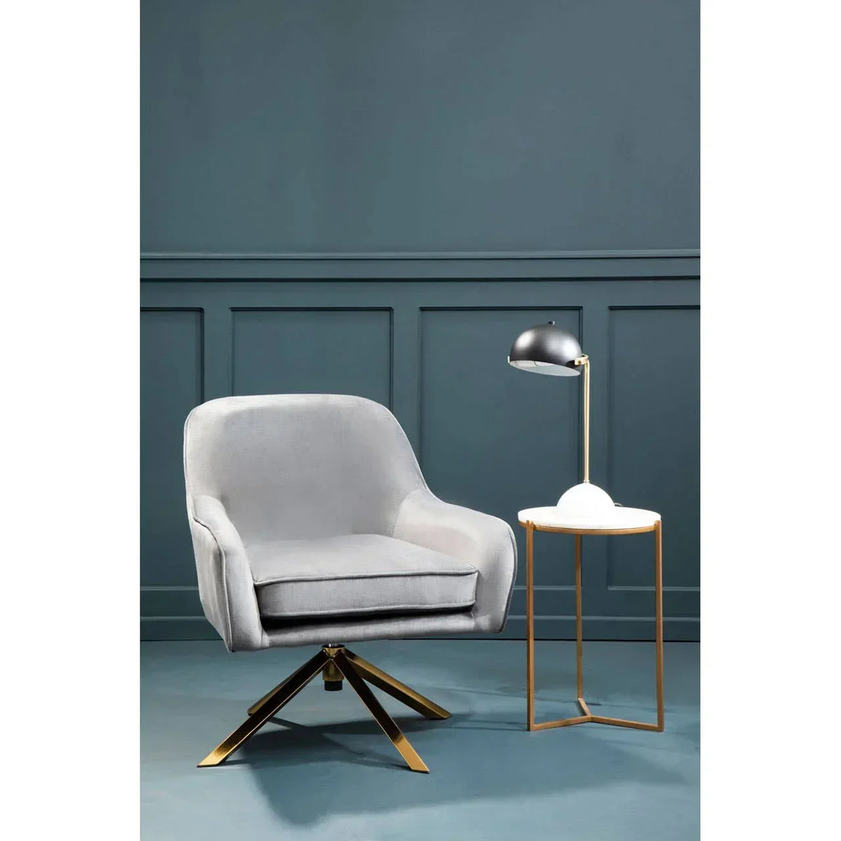 Grey swivel accent chair with gold legs beside marble top side table and desk lamp