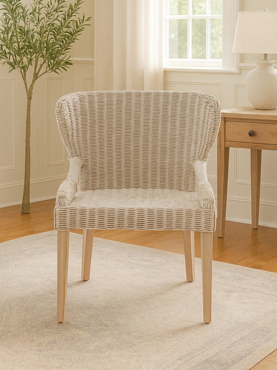 Wicker chair with a curved backrest and light-colored upholstery, featuring wooden legs, placed on a light rug in a bright room with natural light and a small plant in the background