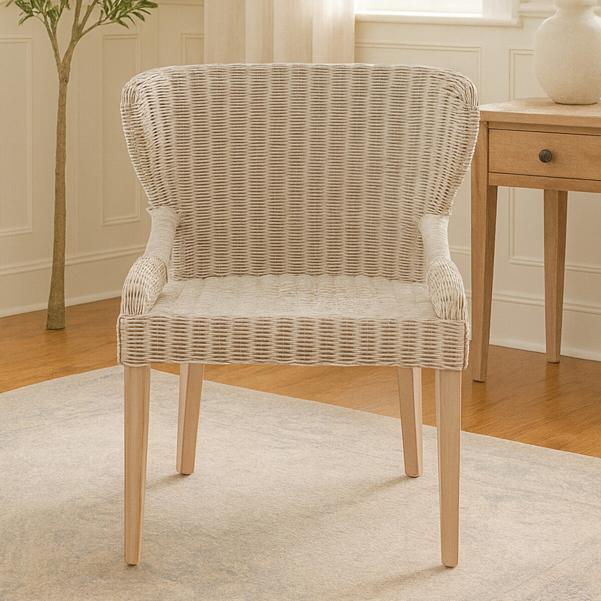Wicker chair with a curved backrest and armrests, featuring a light beige woven pattern, supported by natural wood legs, placed on a light-colored rug in a cozy room setting with a plant and a wooden side table in the background