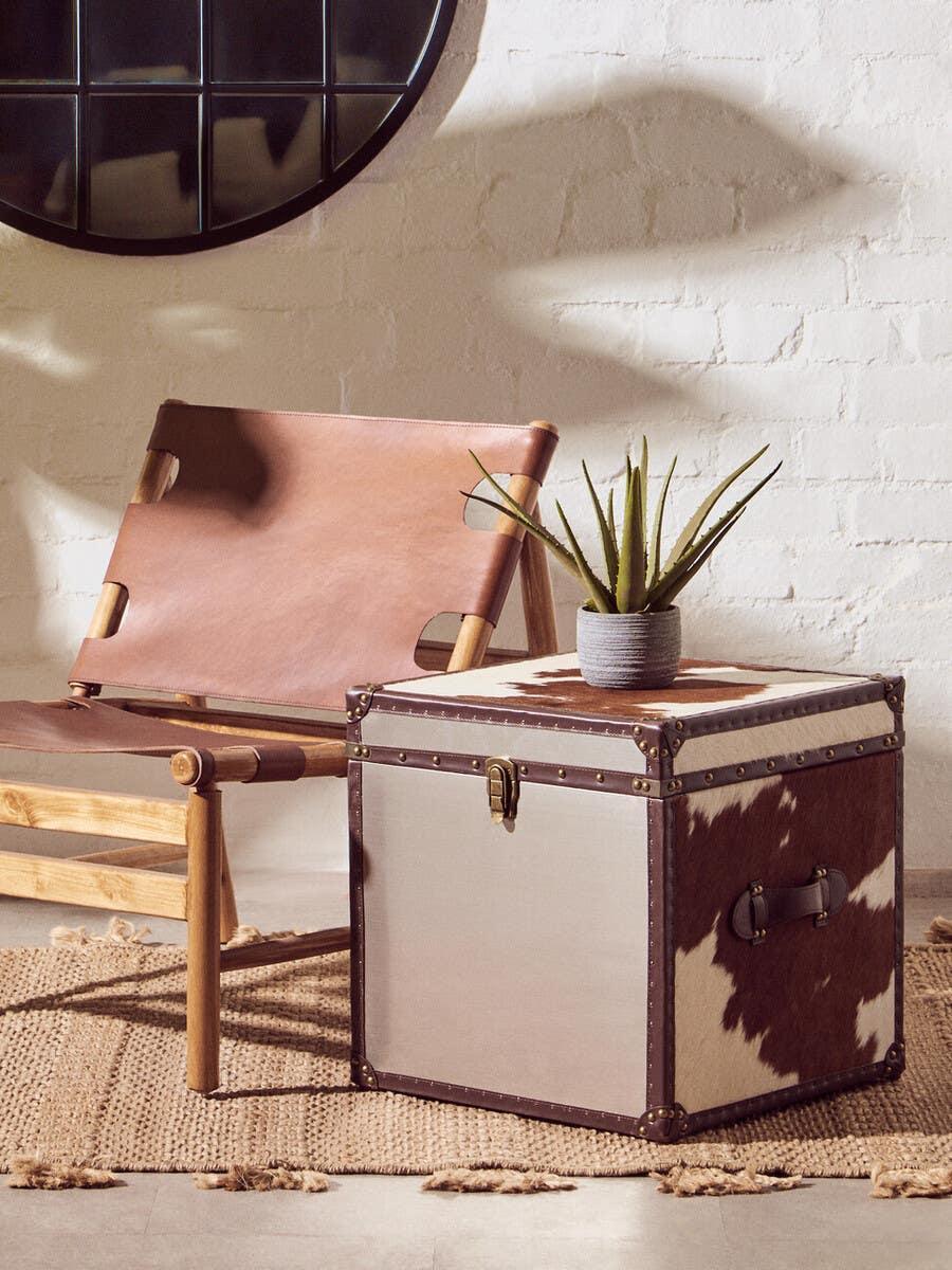 Cowhide storage trunk with brass hardware beside leather accent chair on woven rug