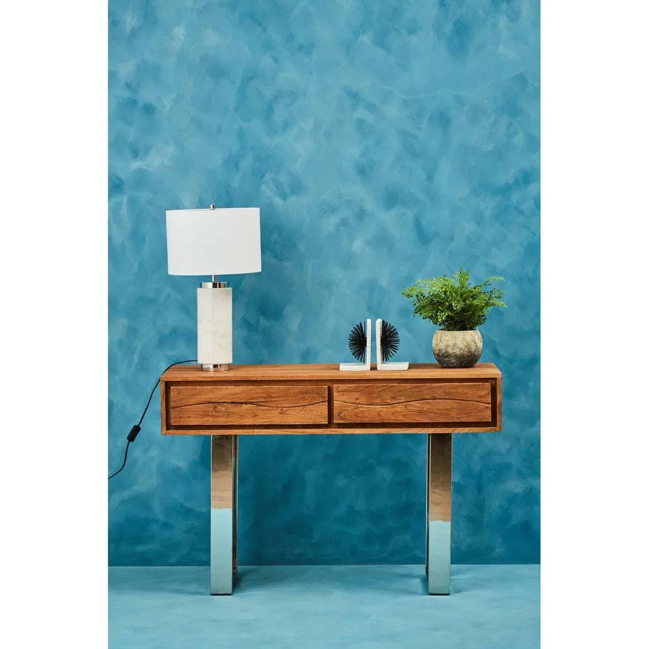 Wood console table with chrome legs, lamp, decor books and potted plant against blue wall