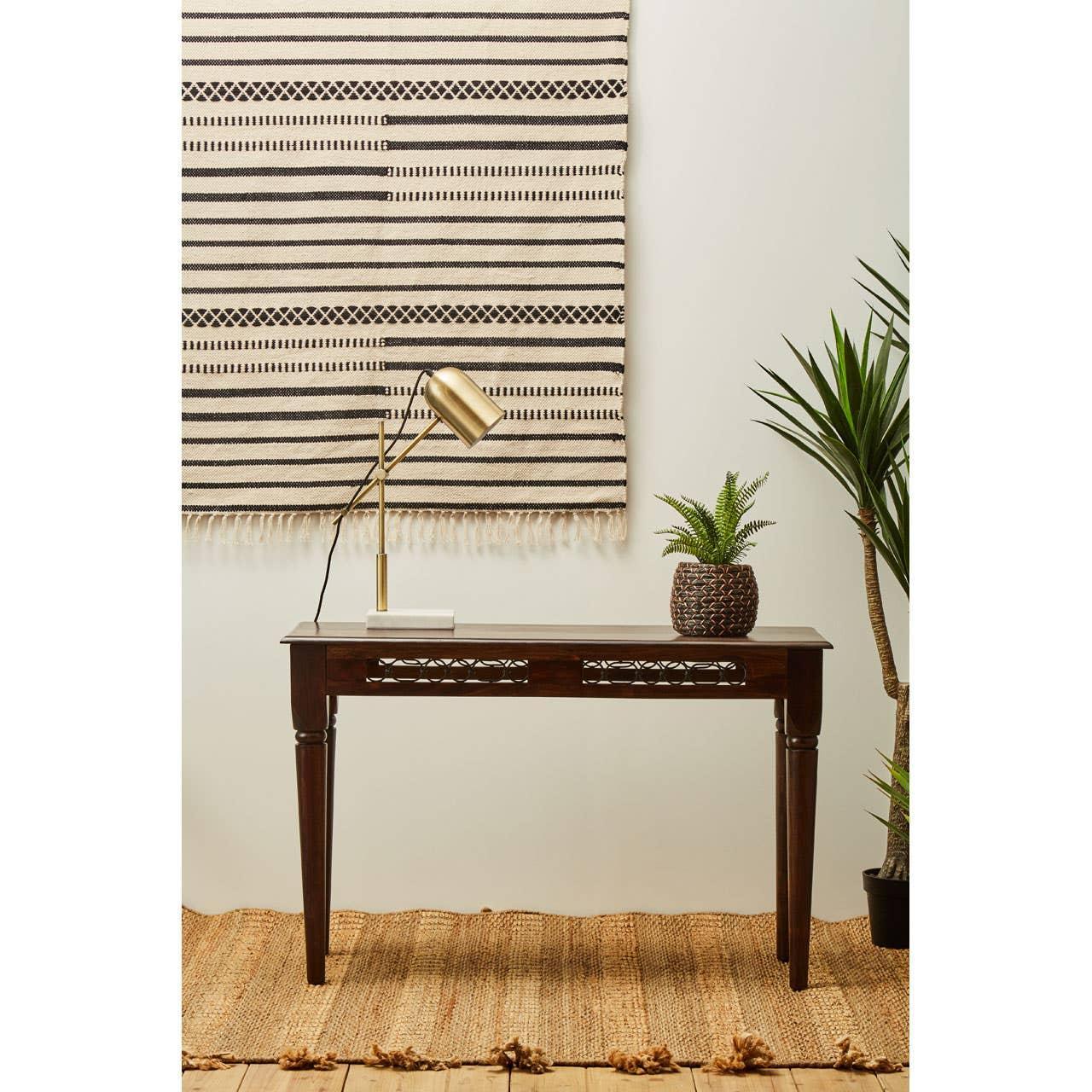 Dark wood console table with brass desk lamp and potted plant beside woven wall hanging