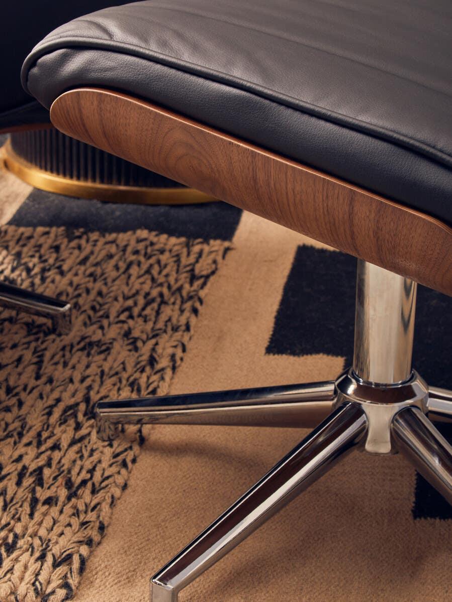 Closeup of black leather footstool with walnut wood trim and chrome base on woven rug