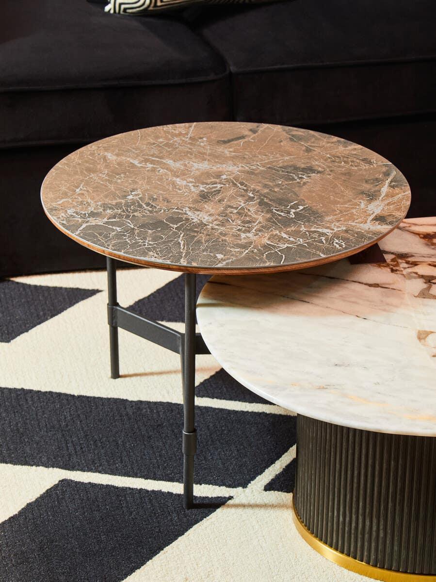 Round brown marble coffee table with black metal legs beside a white marble table on rug