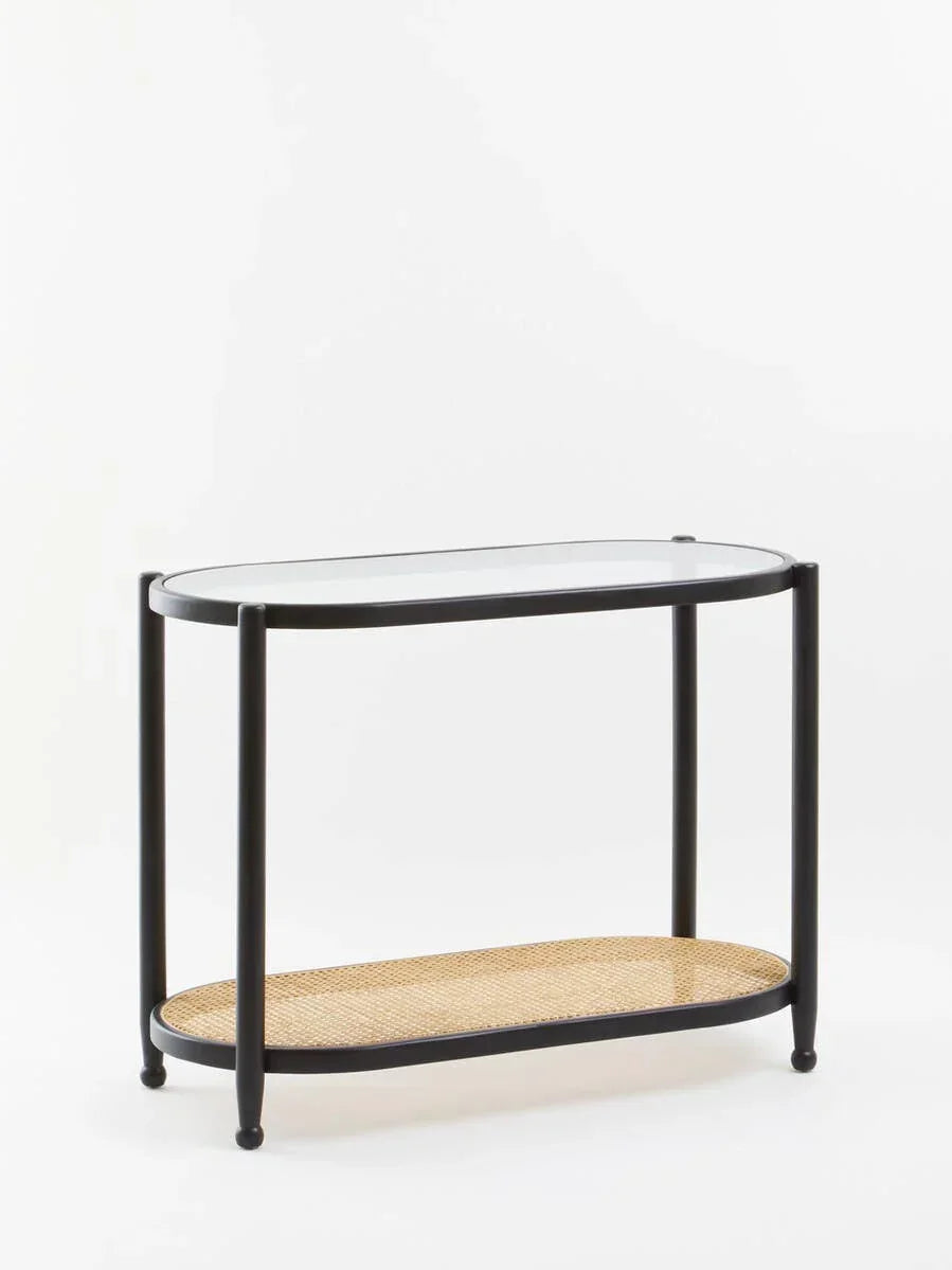 Black metal console table with glass top and rattan shelf on white background