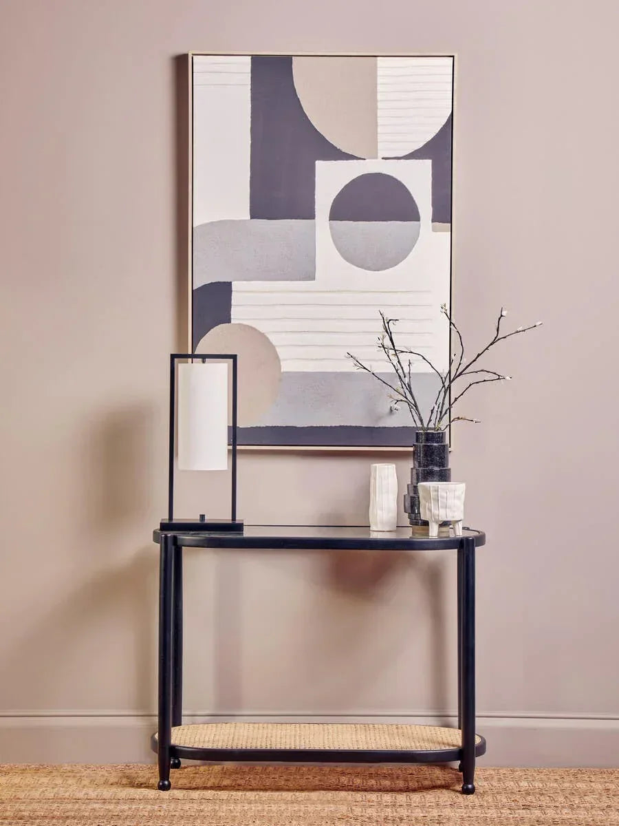 Black console table with rattan shelf, modern lamp, vases and wall art against neutral wall
