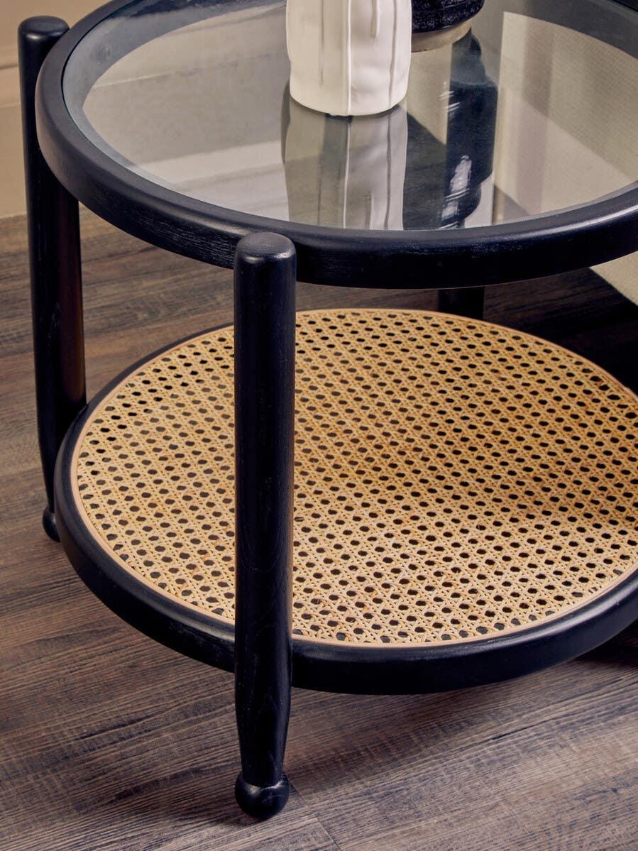 Round glass coffee table with black frame and woven rattan shelf on wood floor