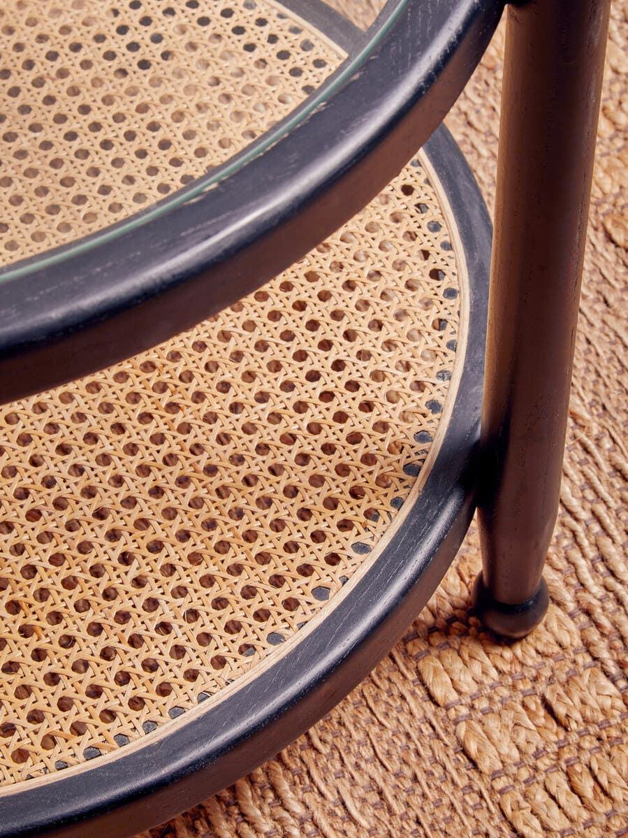 Round rattan side table with black frame and woven shelf on textured rug