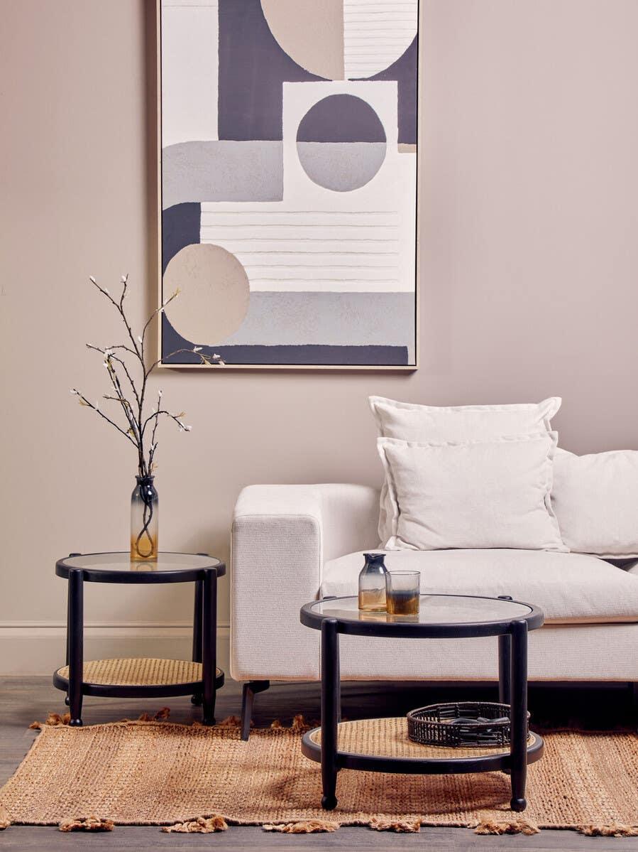 Round black glass coffee tables with woven shelf beside cream sofa and abstract wall art