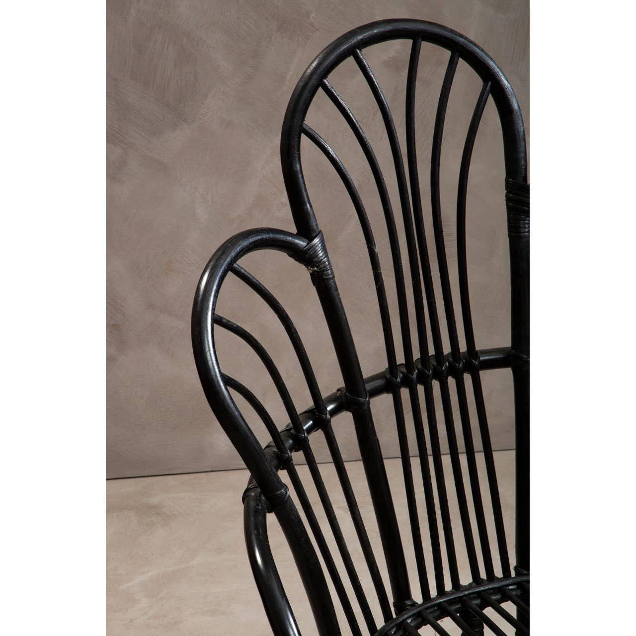 A close-up view of a black metal chair featuring a scalloped back design, with natural rattan detailing. The chair has slender vertical slats and a smooth finish, set against a neutral textured background