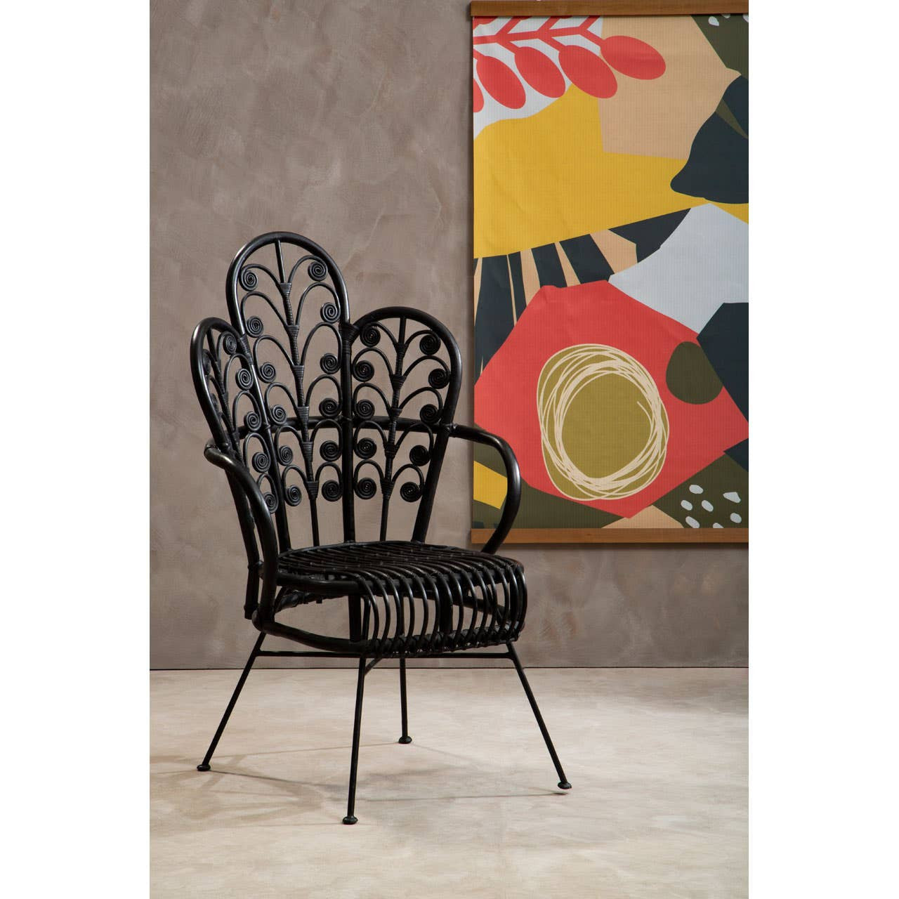 A black rattan chair with a scalloped back design, featuring intricate leaf patterns. The chair has slender metal legs and is positioned against a textured gray wall, with a colorful abstract artwork in the background