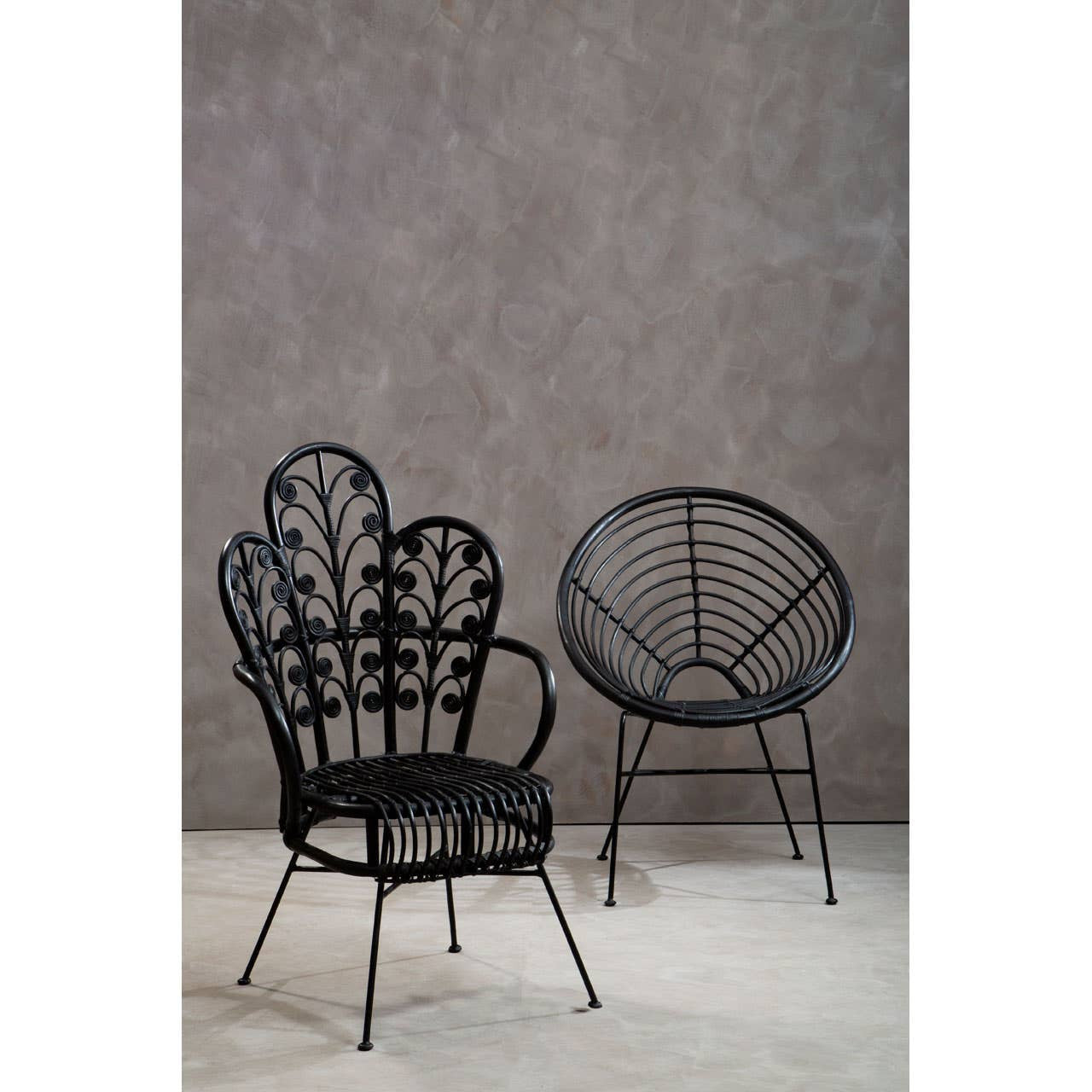 A black natural rattan chair with a scalloped back design is positioned next to a round, open-back chair. The background features a textured gray wall, and the floor is a light, neutral color. The chairs have slender legs and a contemporary aesthetic