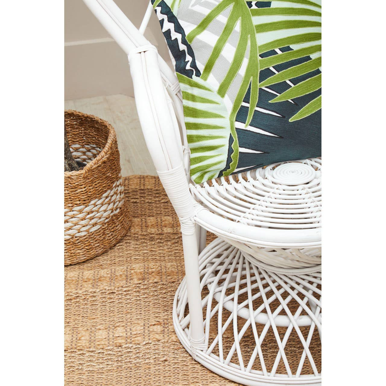Curved chair made of natural rattan in grey and white, featuring a woven design and a round base. A green plant is visible in the background, along with a textured woven basket and a light-colored rug