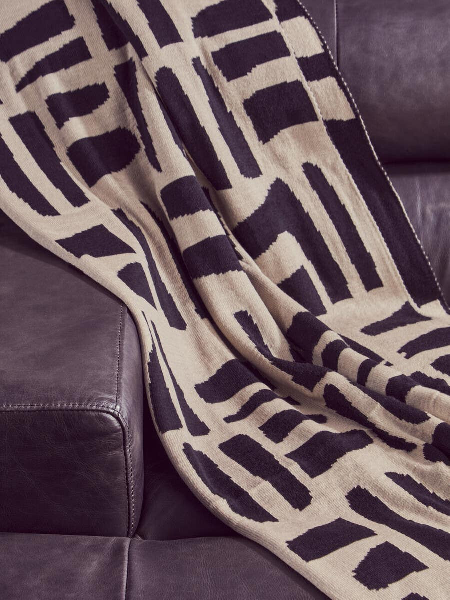 A close-up view of a patterned throw blanket draped over a dark leather sofa, featuring geometric designs in black and beige. The texture of the leather is visible, highlighting the luxurious feel of the chaise