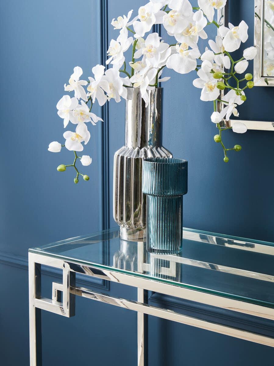 Glass console table with chrome frame, silver vase with white artificial flowers, blue wall