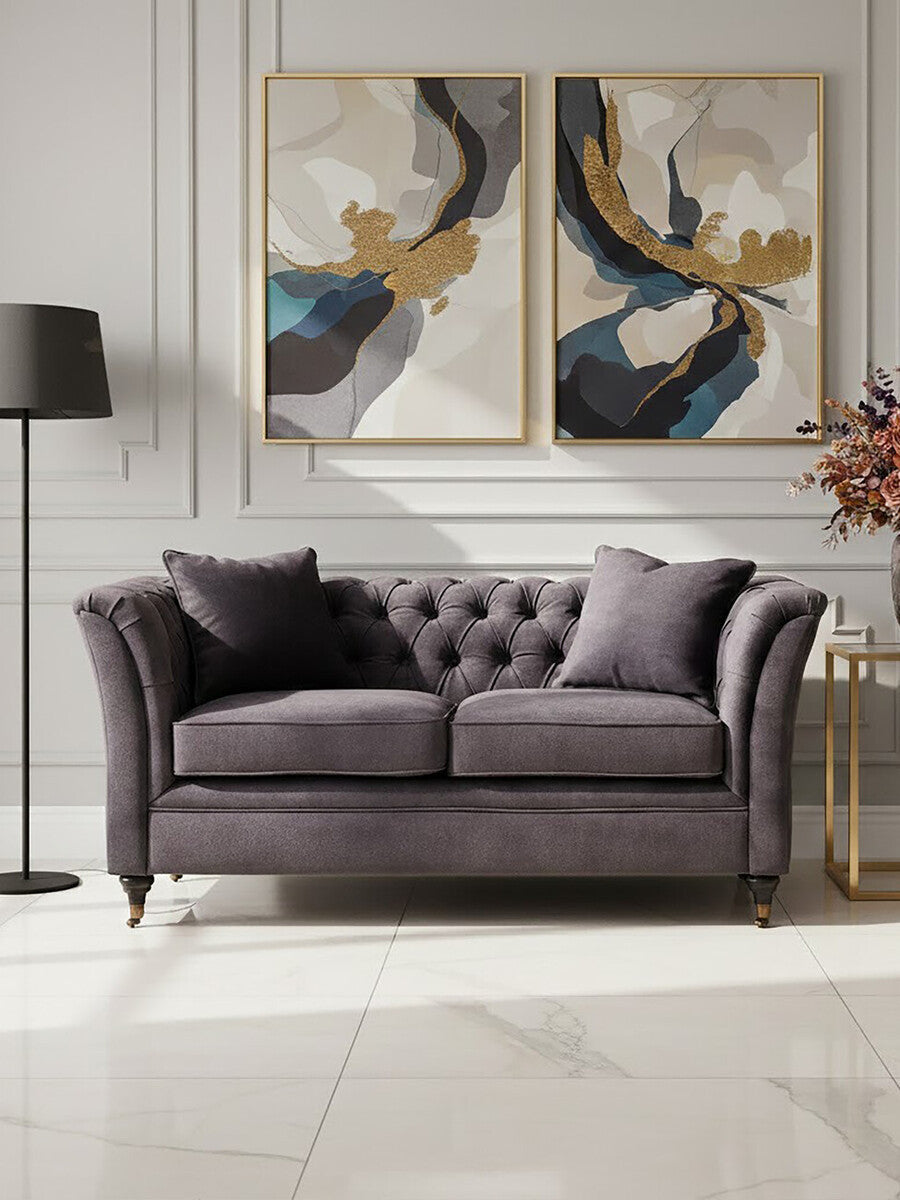 Two-seat grey velvet sofa with tufted backrest and plush cushions, positioned in a stylish living room setting. The sofa is complemented by two decorative pillows and is placed against a wall featuring two abstract art pieces. A modern floor lamp stands nearby, and a small side table holds a vase with flowers. The flooring is light-colored, enhancing the overall elegance of the space