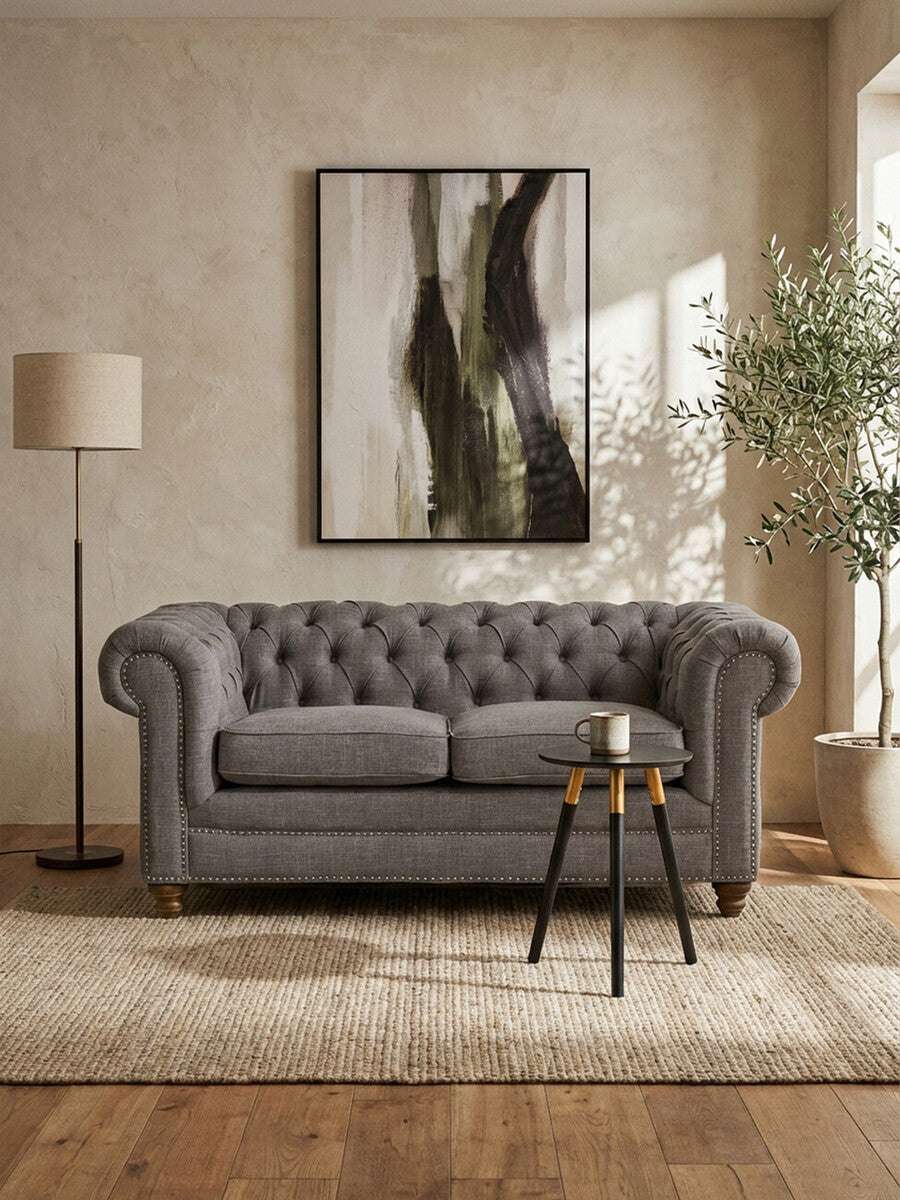 Two-seat grey linen sofa with tufted backrest, placed in a bright room featuring a large abstract wall art piece, a standing lamp, and a small round side table with decorative items, surrounded by a light-colored rug and a potted plant