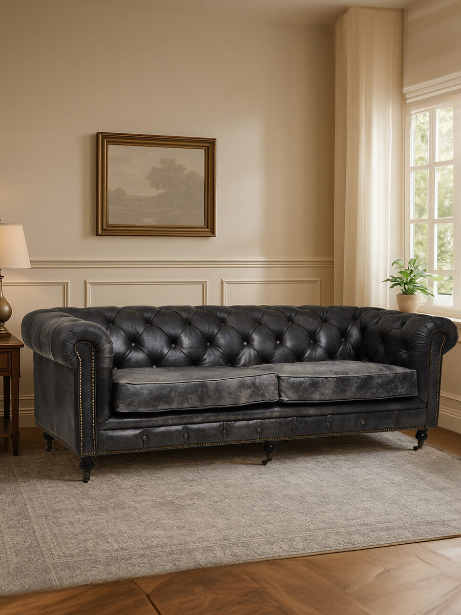 Antique black Chesterfield leather sofa with three seats, featuring tufted backrest and armrests, set against a light-colored wall with a framed artwork and a lamp on a side table, complemented by a soft area rug on the floor