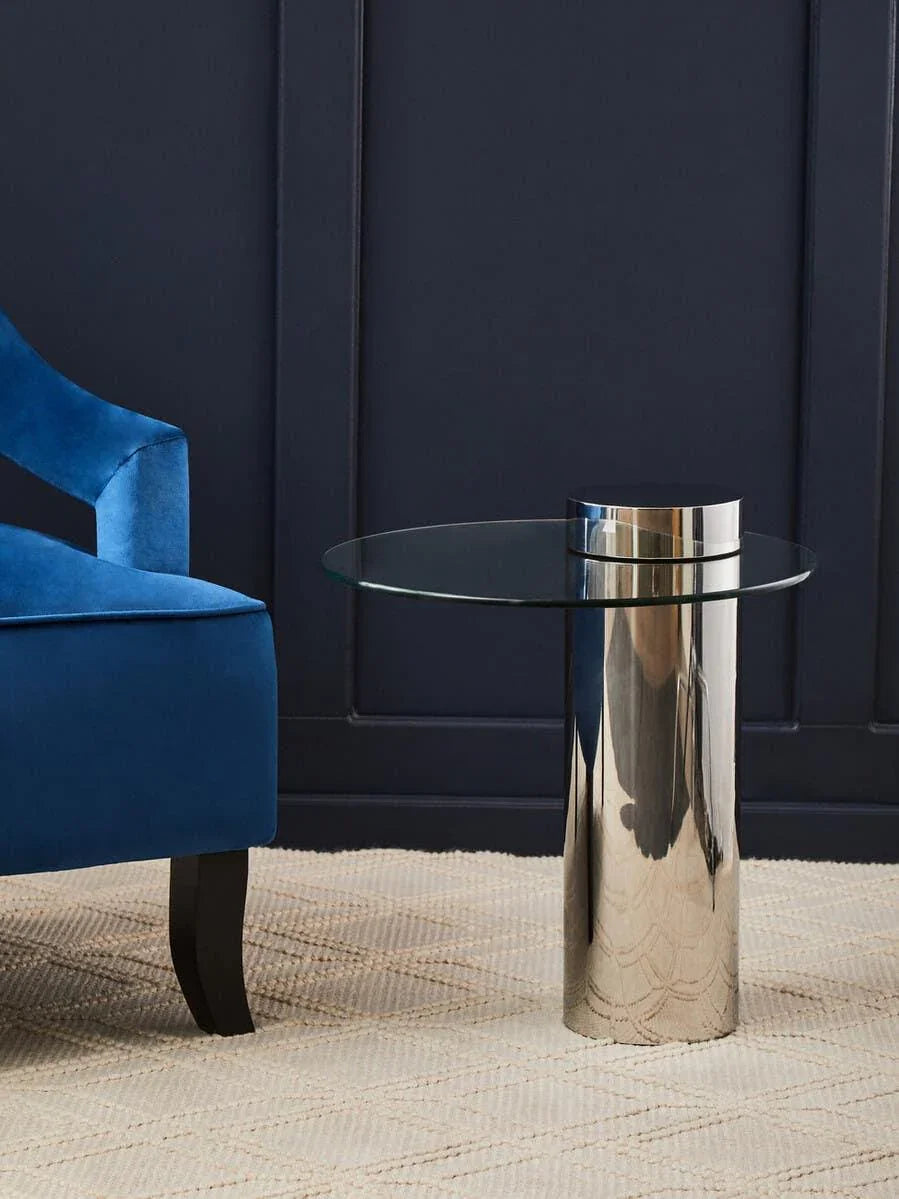 Glass top side table with chrome cylindrical base beside blue velvet armchair