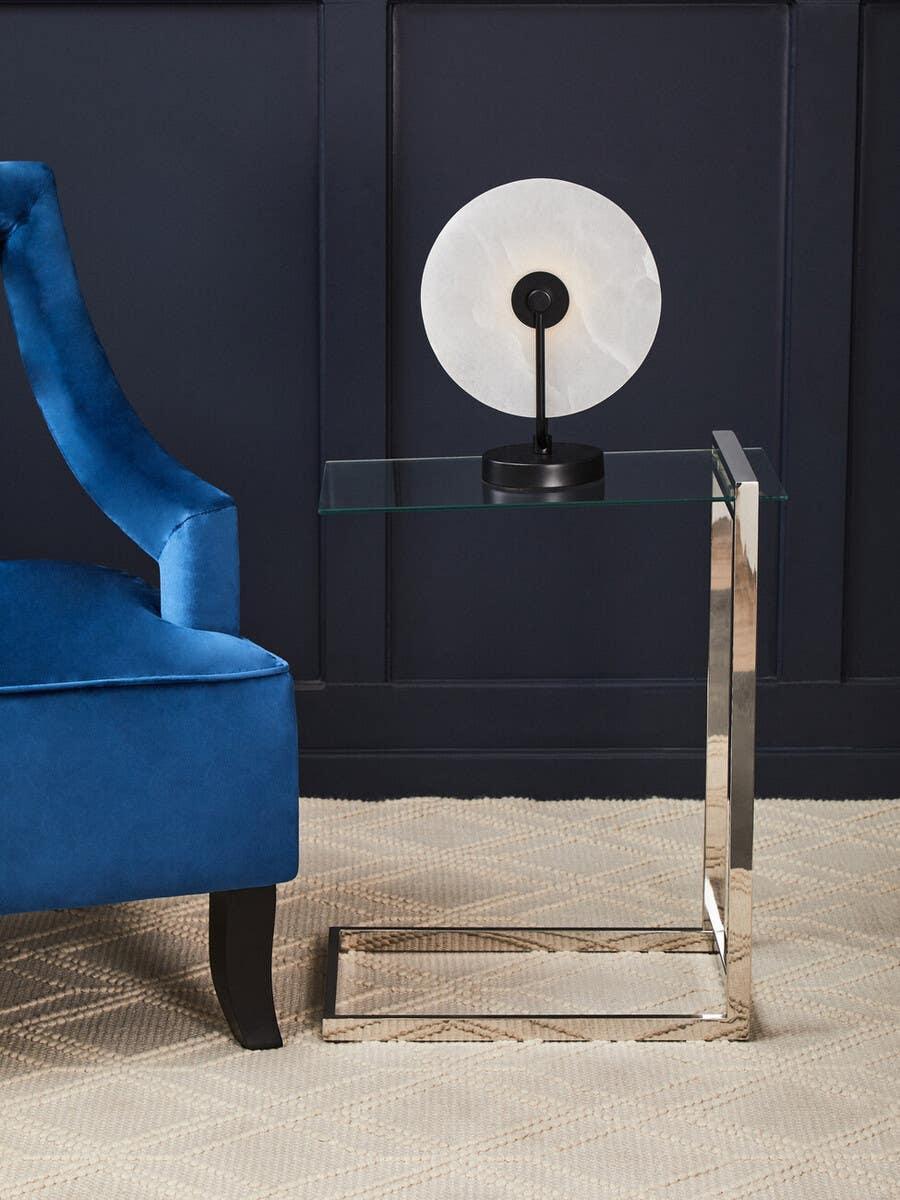 Glass and chrome side table beside blue velvet armchair, decorative lamp on top