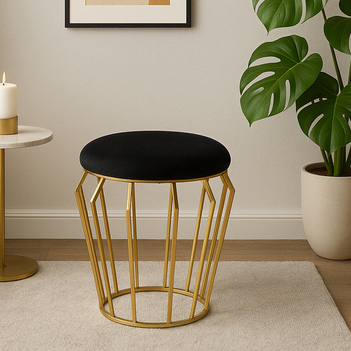 A black velvet stool with a round top and a gold metal wire frame, placed on a light-colored floor in a modern living space. A green plant and a small side table are visible in the background