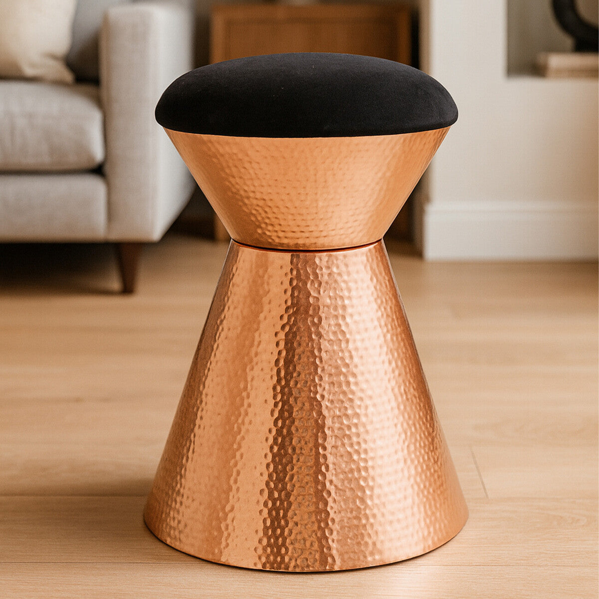 A stylish stool featuring a black velvet top and a hammered copper finish base, designed with a modern, tapered silhouette. The stool is placed on a light wooden floor, with a soft, neutral-toned sofa visible in the background