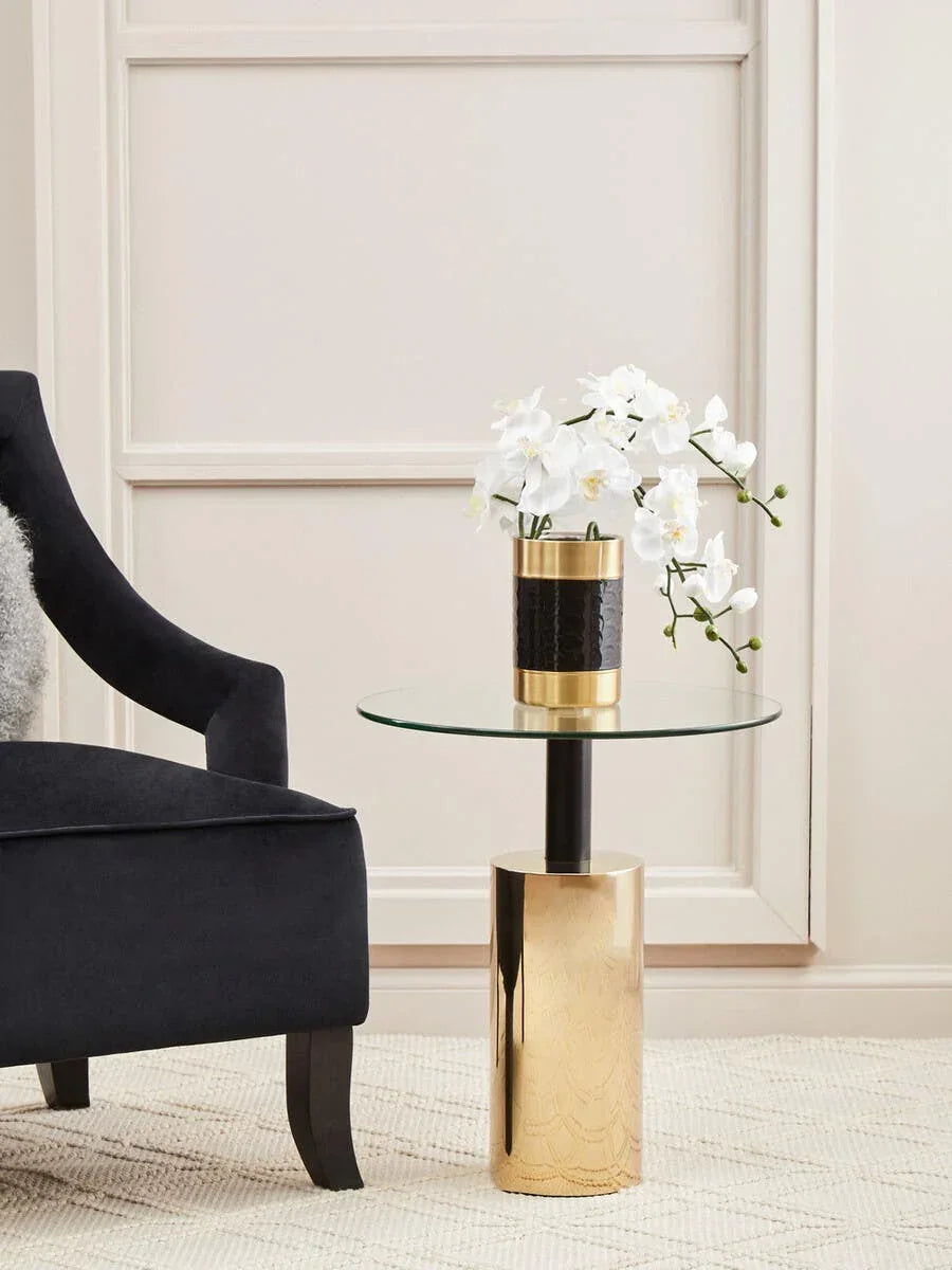 Glass top side table with gold metal base beside black armchair and white orchid vase