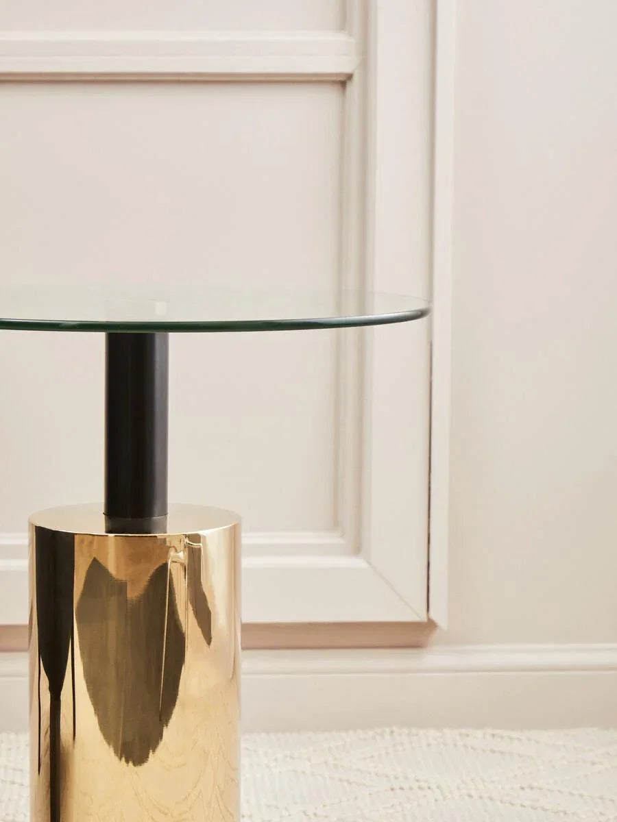 Round glass top side table with gold metal base and black stem against cream wall