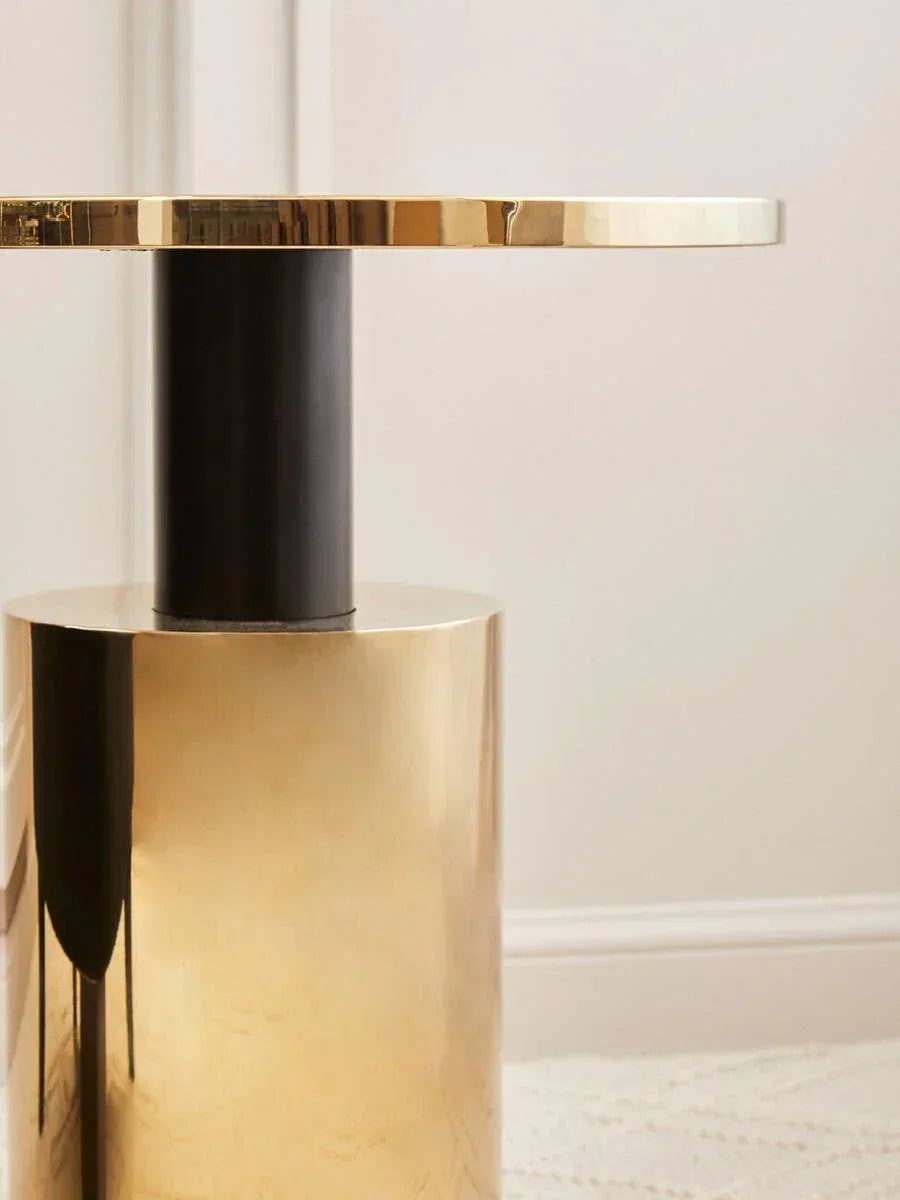 Gold metal side table with cylindrical base and round top against neutral wall