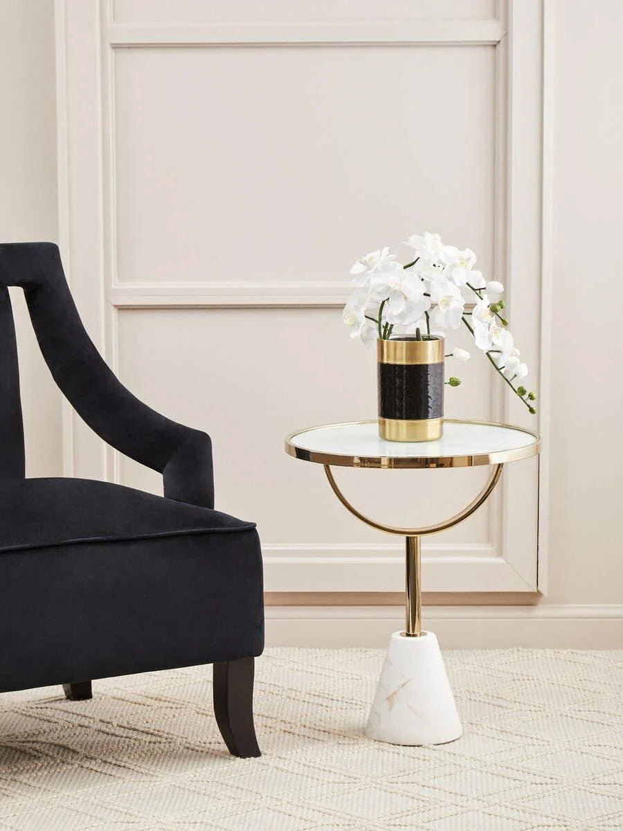 Glass top side table with gold frame and marble base beside black velvet armchair