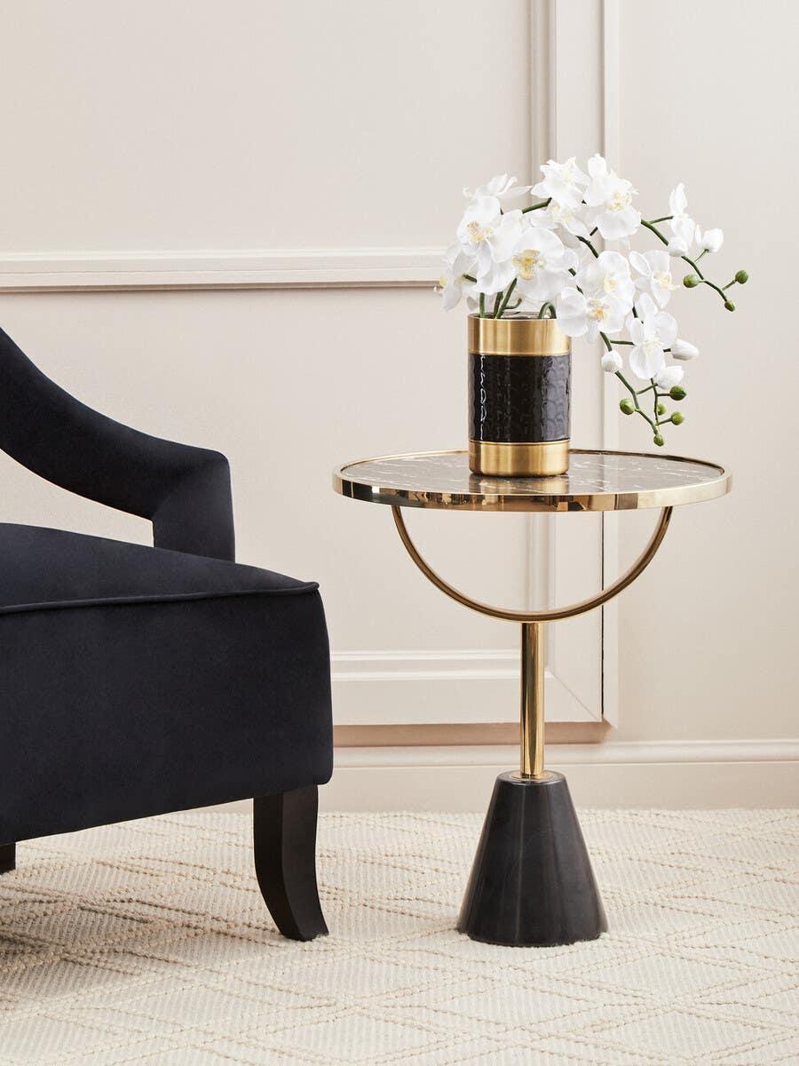 Gold and black side table with white orchid vase beside dark fabric armchair on cream rug