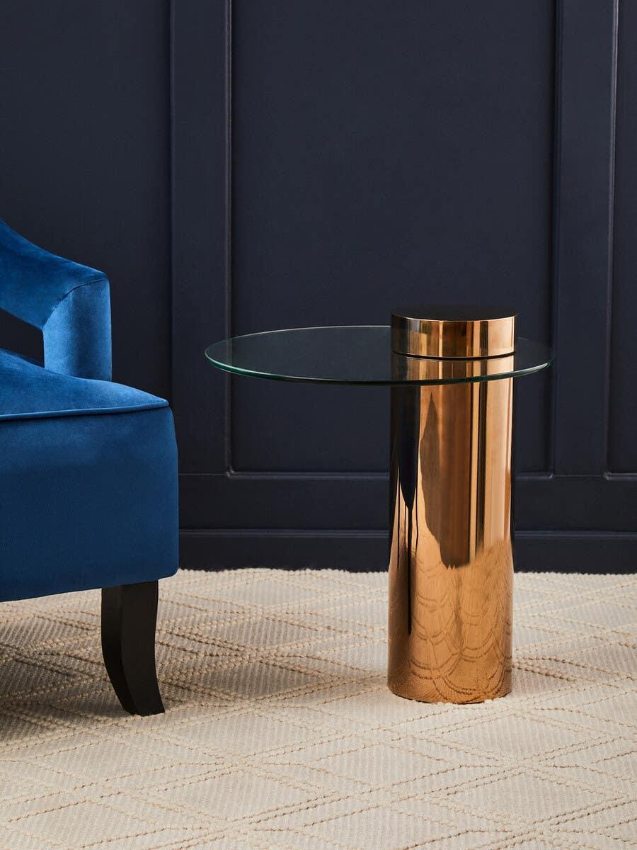 Glass side table with bronze metallic base beside blue velvet armchair on cream rug