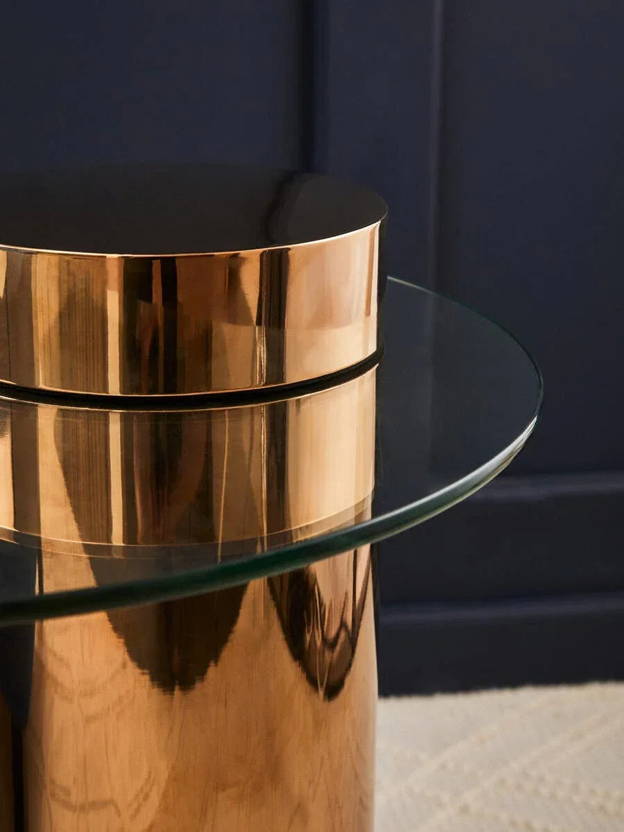 Round glass side table with gold metallic base against dark wall