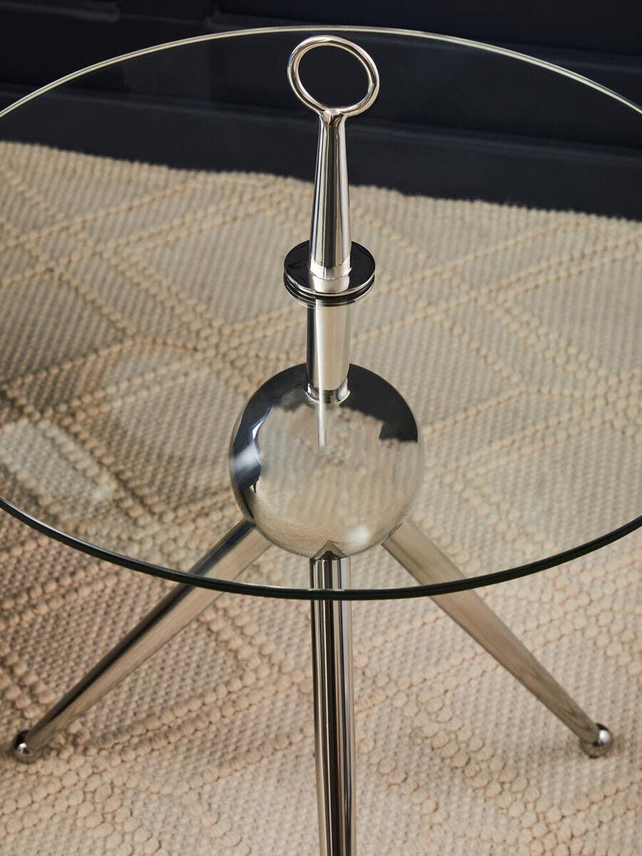 Round glass side table with chrome tripod legs and decorative handle detail on beige rug