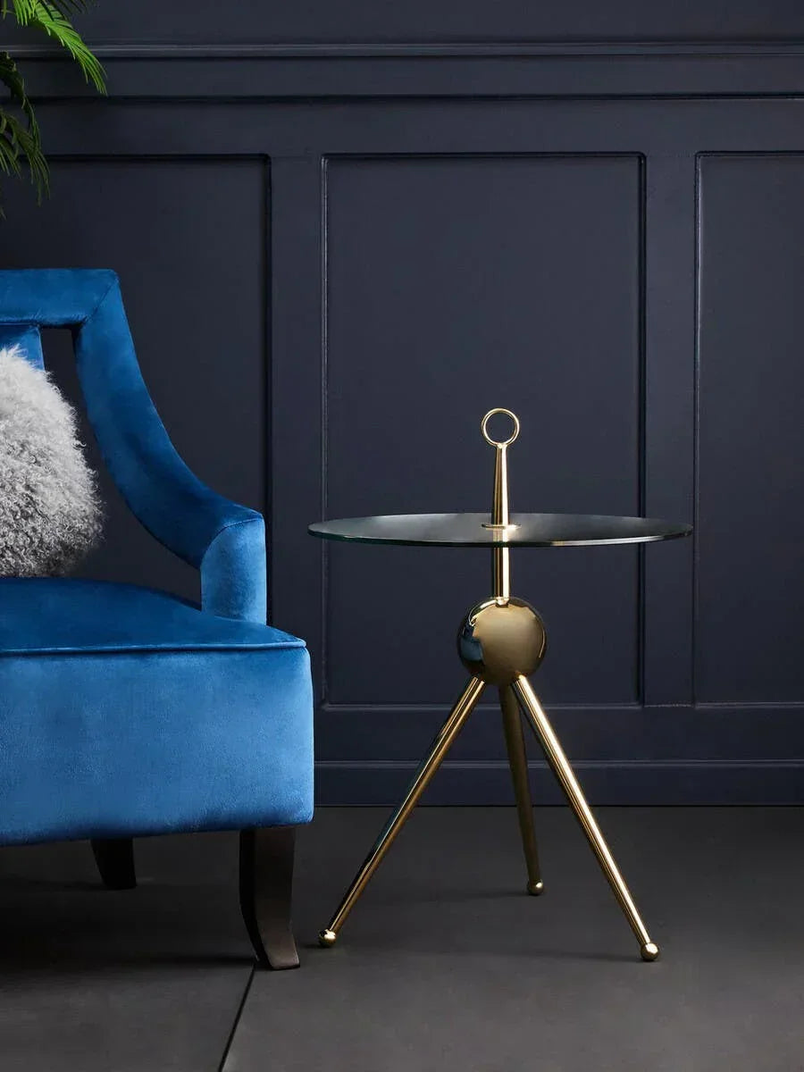 Glass top side table with gold tripod legs beside blue velvet armchair