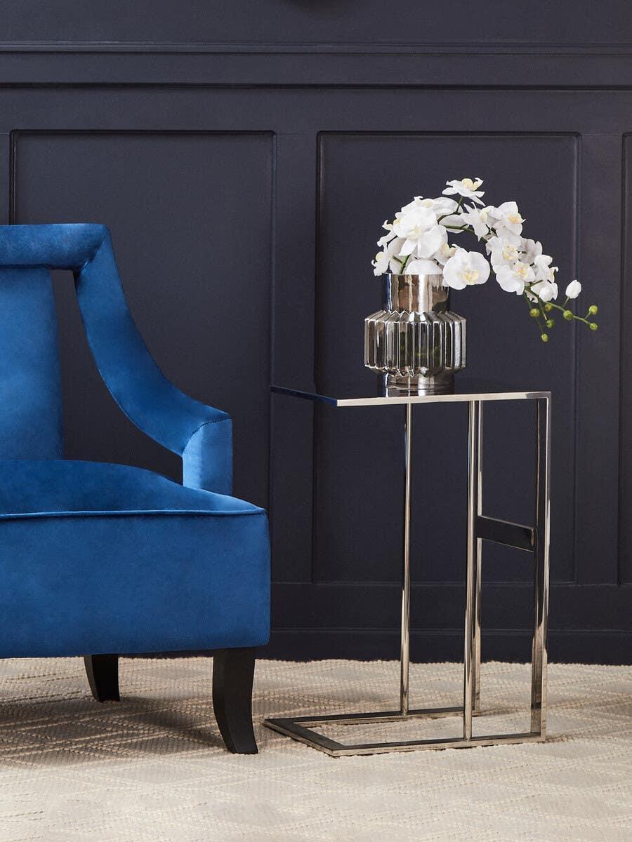 Chrome side table with glass vase and white flowers beside blue velvet accent chair
