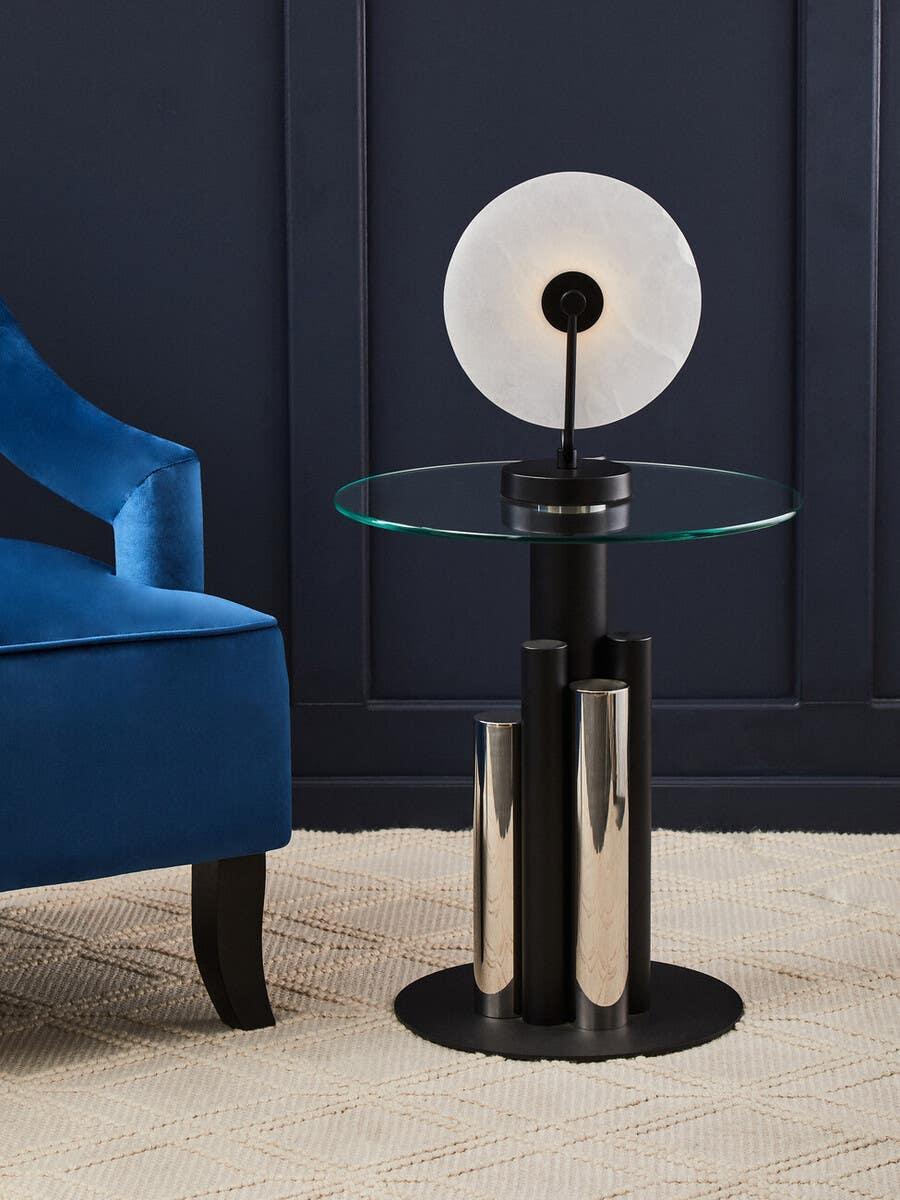 Glass top side table with chrome cylindrical legs and modern lamp against dark wall