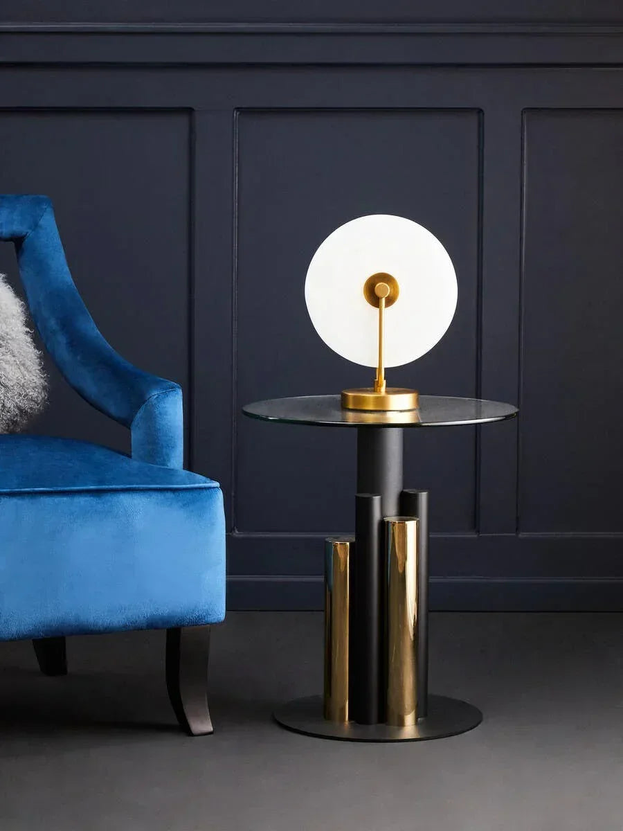 Modern glass and brass side table with round lamp beside blue velvet armchair