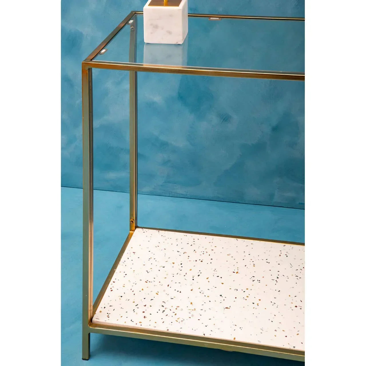 Gold metal side table with glass top and speckled terrazzo lower shelf against blue wall