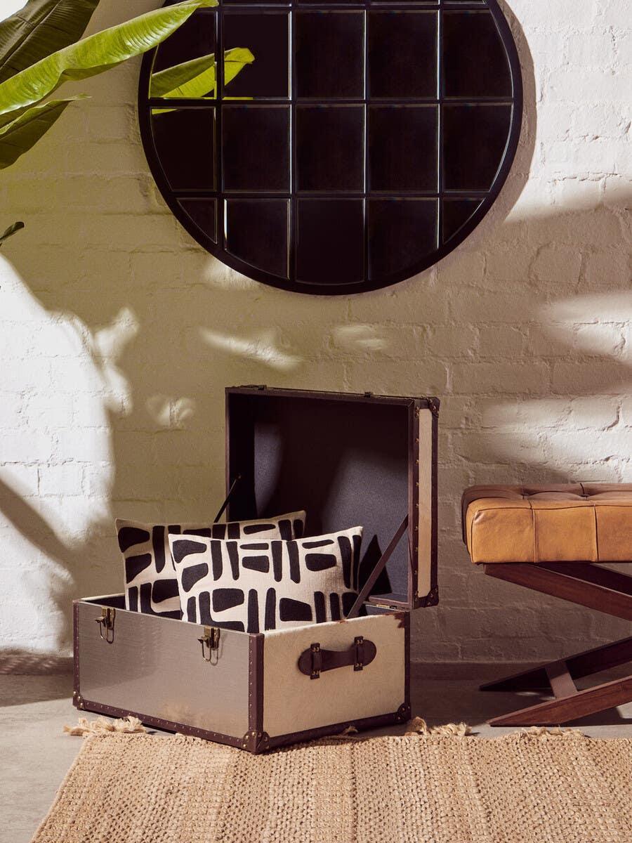 Storage trunk with black and white patterned cushions beside tan leather bench on jute rug