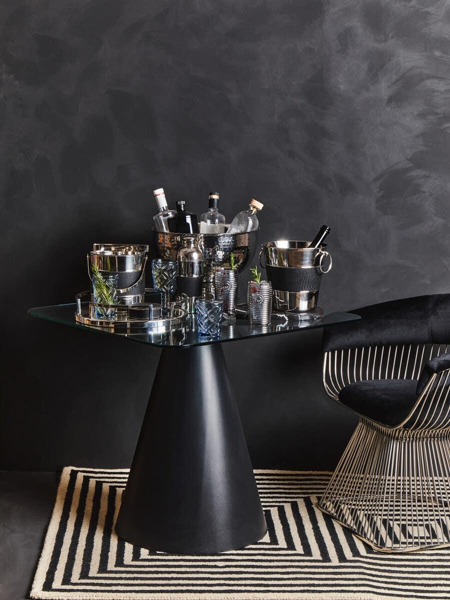 Glass dining table with black cone base, metal chair, and drinks accessories on striped rug