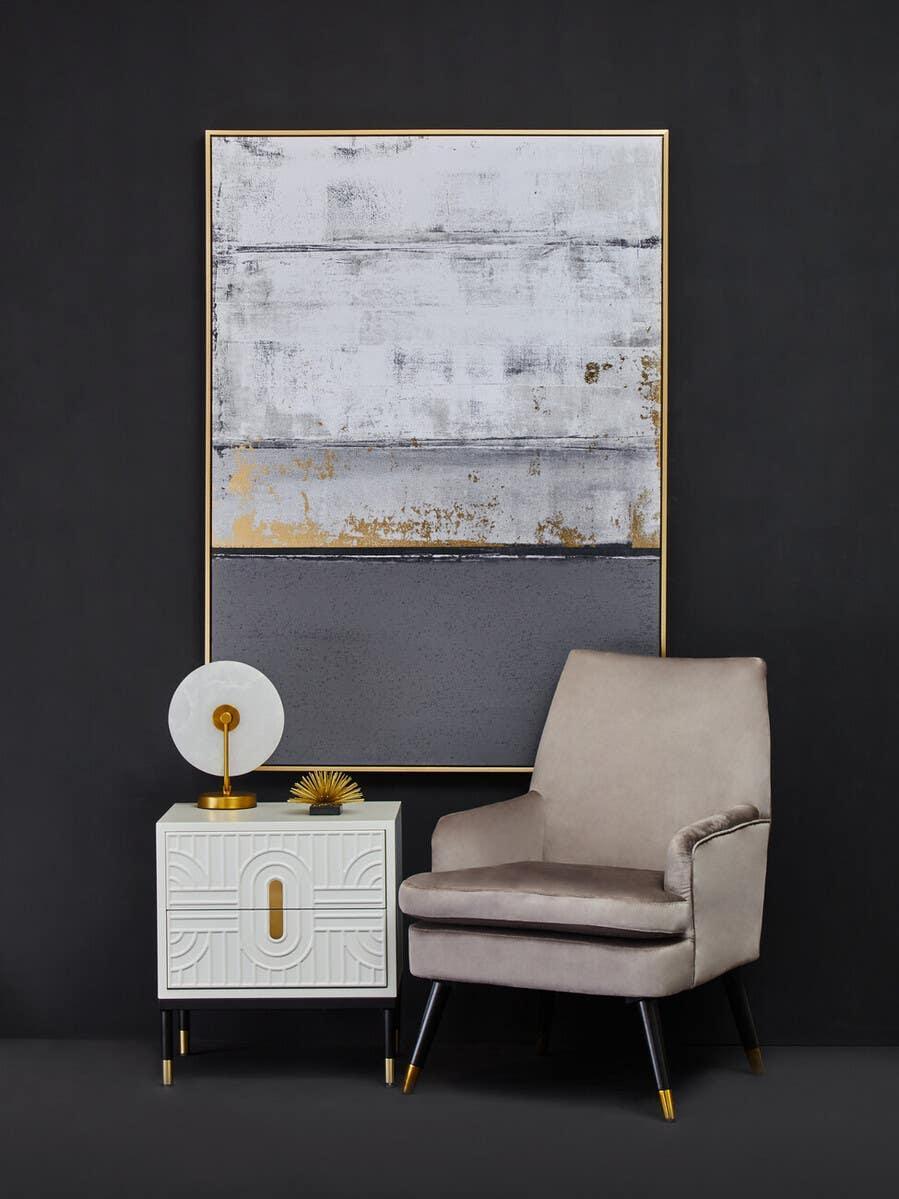 Grey velvet accent chair with gold-tipped legs beside modern white bedside table and large abstract art